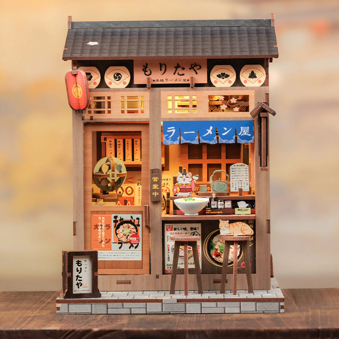From the iconic noren curtain swaying above the window to the finely detailed counter lined with ramen bowls, rice balls, and sake bottles, every element of the Ramen Sake House DIY book nook kit captures the warm, nostalgic spirit of a cozy late-night eatery tucked away in a quiet Tokyo alley. Soft LED lights bathe the scene in a gentle glow, evoking the hum of conversation, the clink of dishes, and the comforting aroma of a midnight meal.