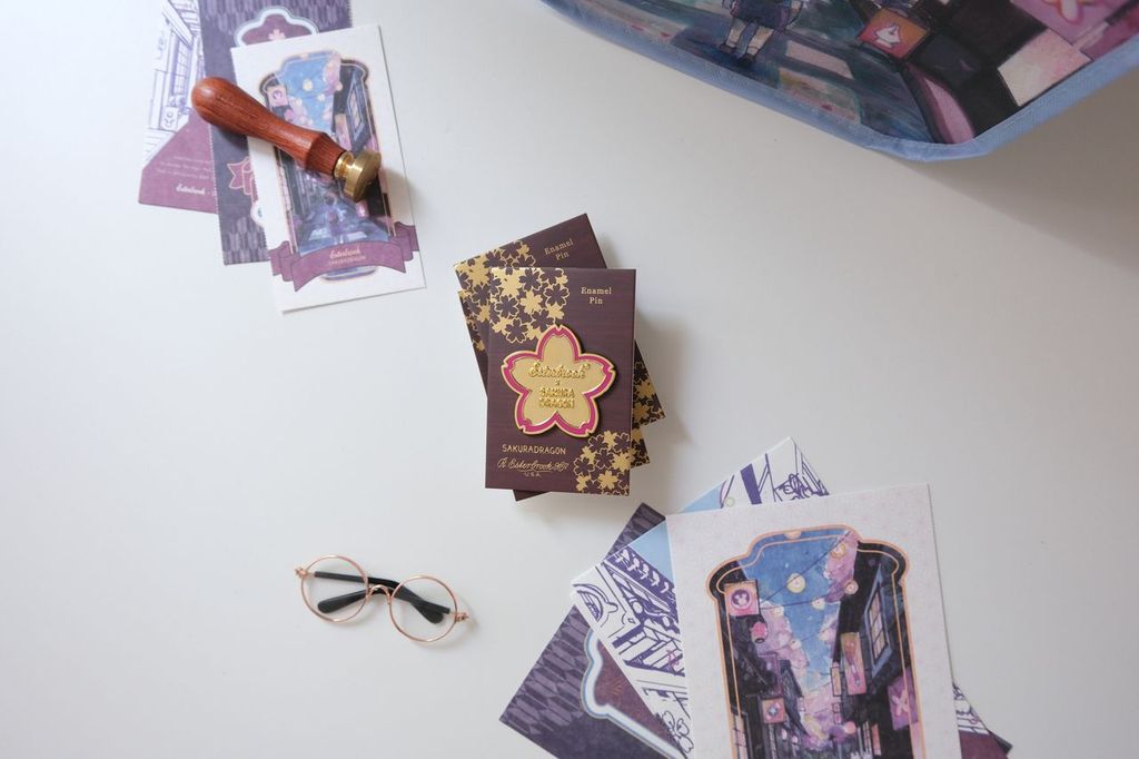 Inspired by the blossoms found throughout Cindy Duong's Sakura Street illustrations, this oversized hard enamel pin features a sakura flower outlined in gold-plated metal. The 5 cm design includes Esterbrook and Sakuradragon branding at its center, creating a collectible emblem of the collaboration.