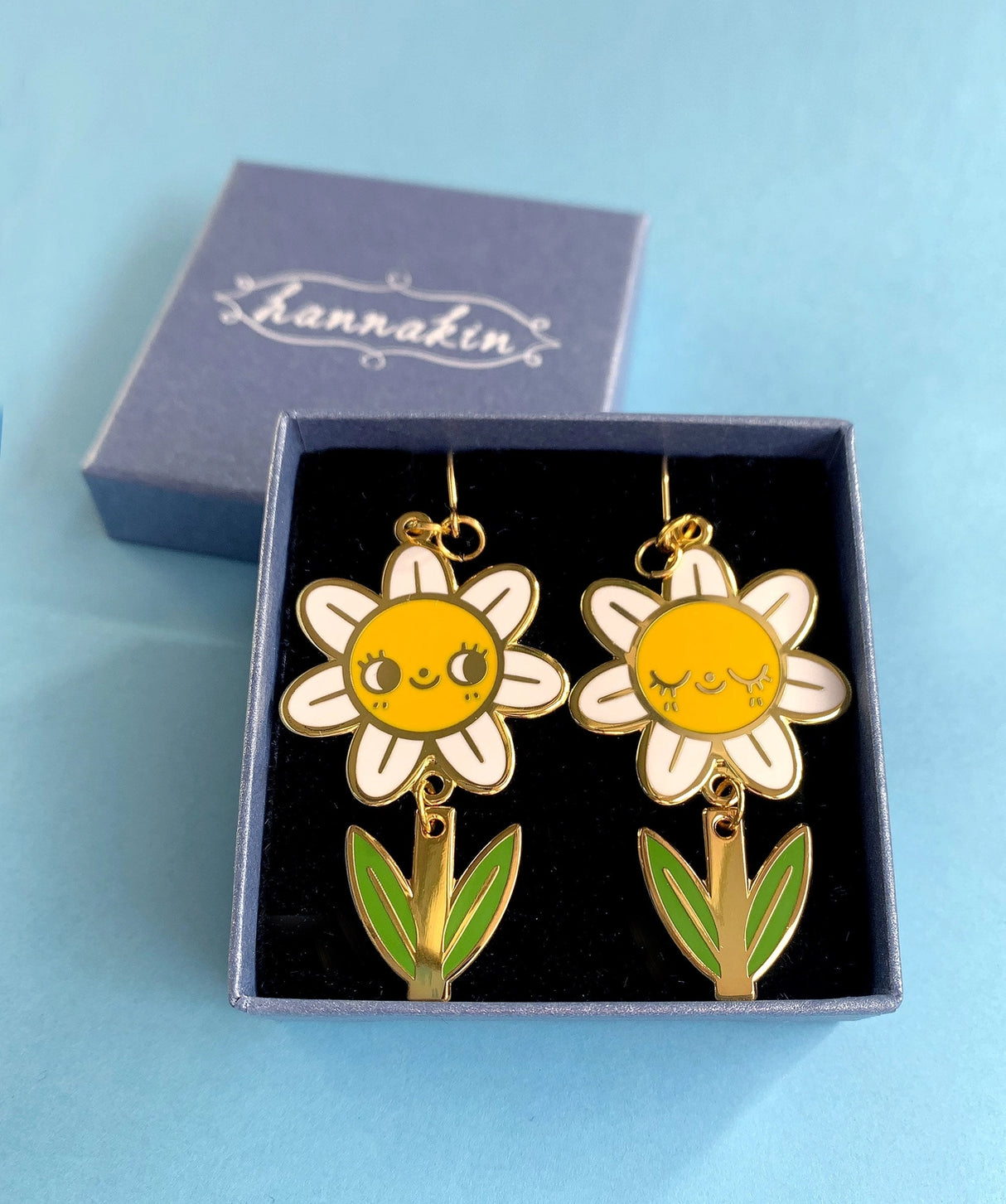A pair of mis-matched happy daisy themed earrings (one one with happy open eyes, and one with happy closed) with separate dangling stems. Made from hard enamel with shiny gold metal finish on high quality Hypo-Allergenic, gold plated, brass based hooks.
Each earring measures approximately 5cm tall x 3cm wide. Earrings come in a Hannakin embossed gift box.