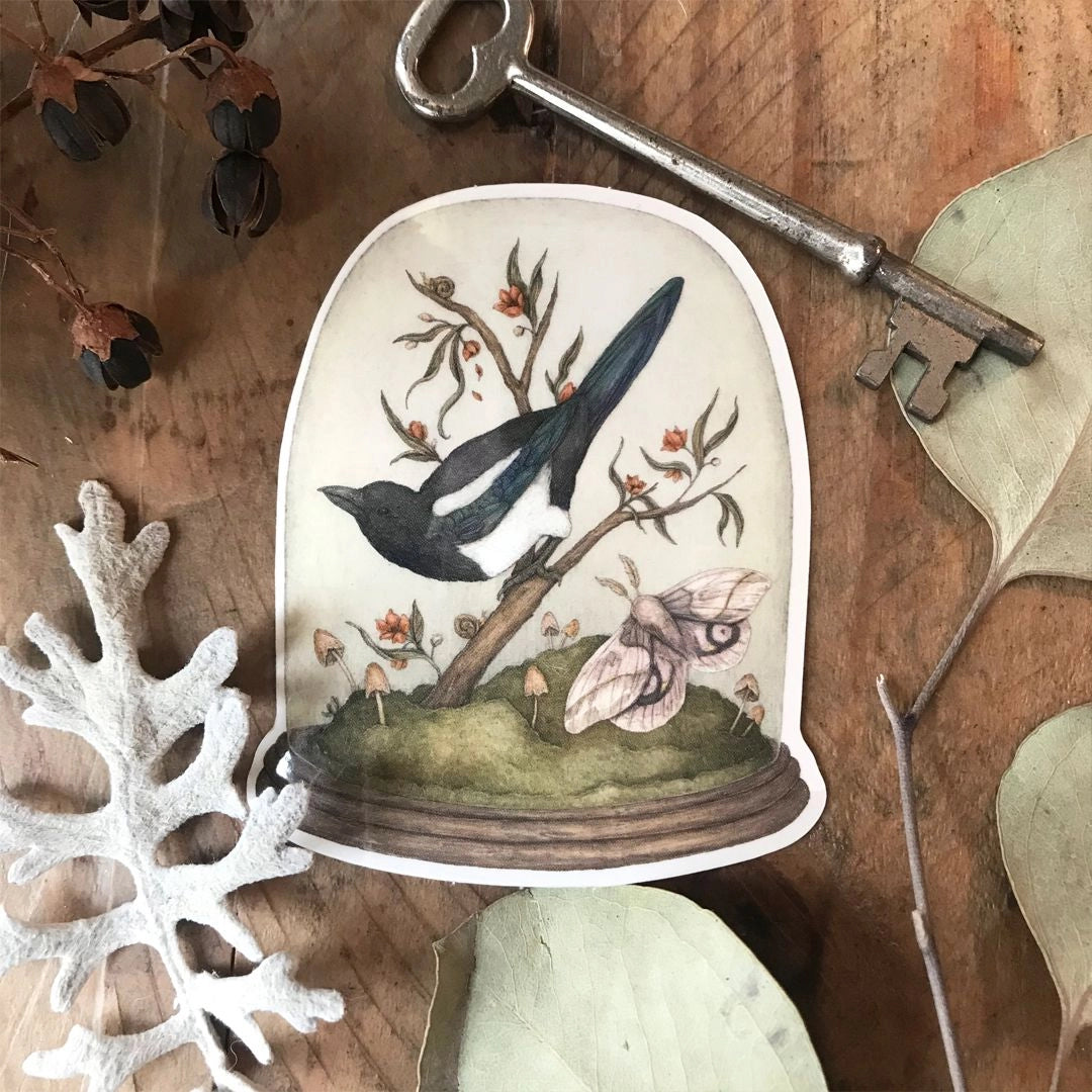 Vinyl semi-transparent sticker of One for Sorrow, an illustration of a magpie and moth under a cloche jar, by Jessica Roux.
The glass of the jar is transparent, but the contents and base of the jar are opaque. Sticker measures 2.6" x 3.1" and is glossy.