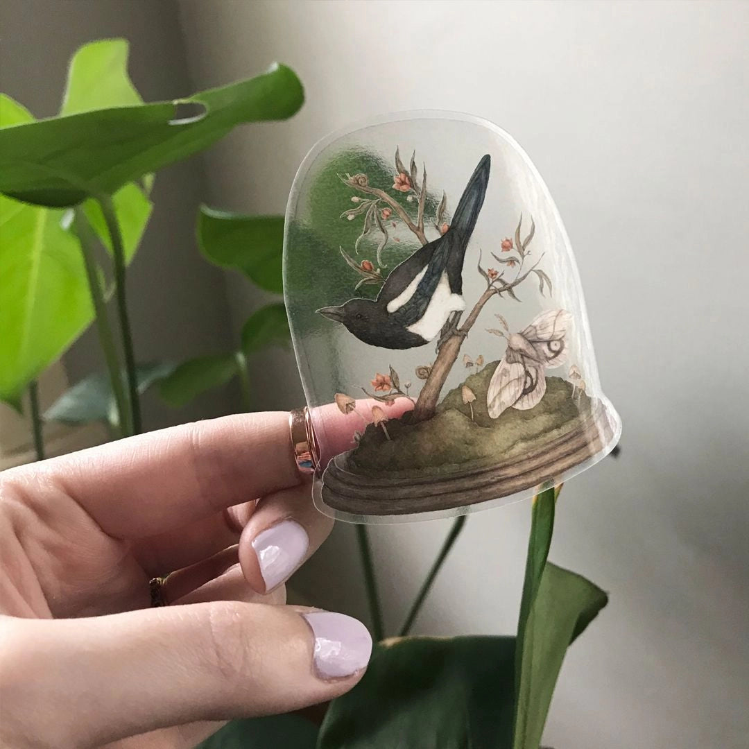 Vinyl semi-transparent sticker of One for Sorrow, an illustration of a magpie and moth under a cloche jar, by Jessica Roux.
The glass of the jar is transparent, but the contents and base of the jar are opaque. Sticker measures 2.6" x 3.1" and is glossy.