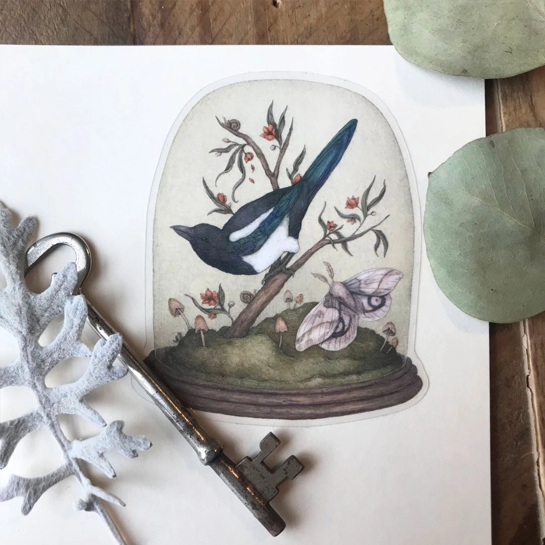 Vinyl semi-transparent sticker of One for Sorrow, an illustration of a magpie and moth under a cloche jar, by Jessica Roux.
The glass of the jar is transparent, but the contents and base of the jar are opaque. Sticker measures 2.6" x 3.1" and is glossy.