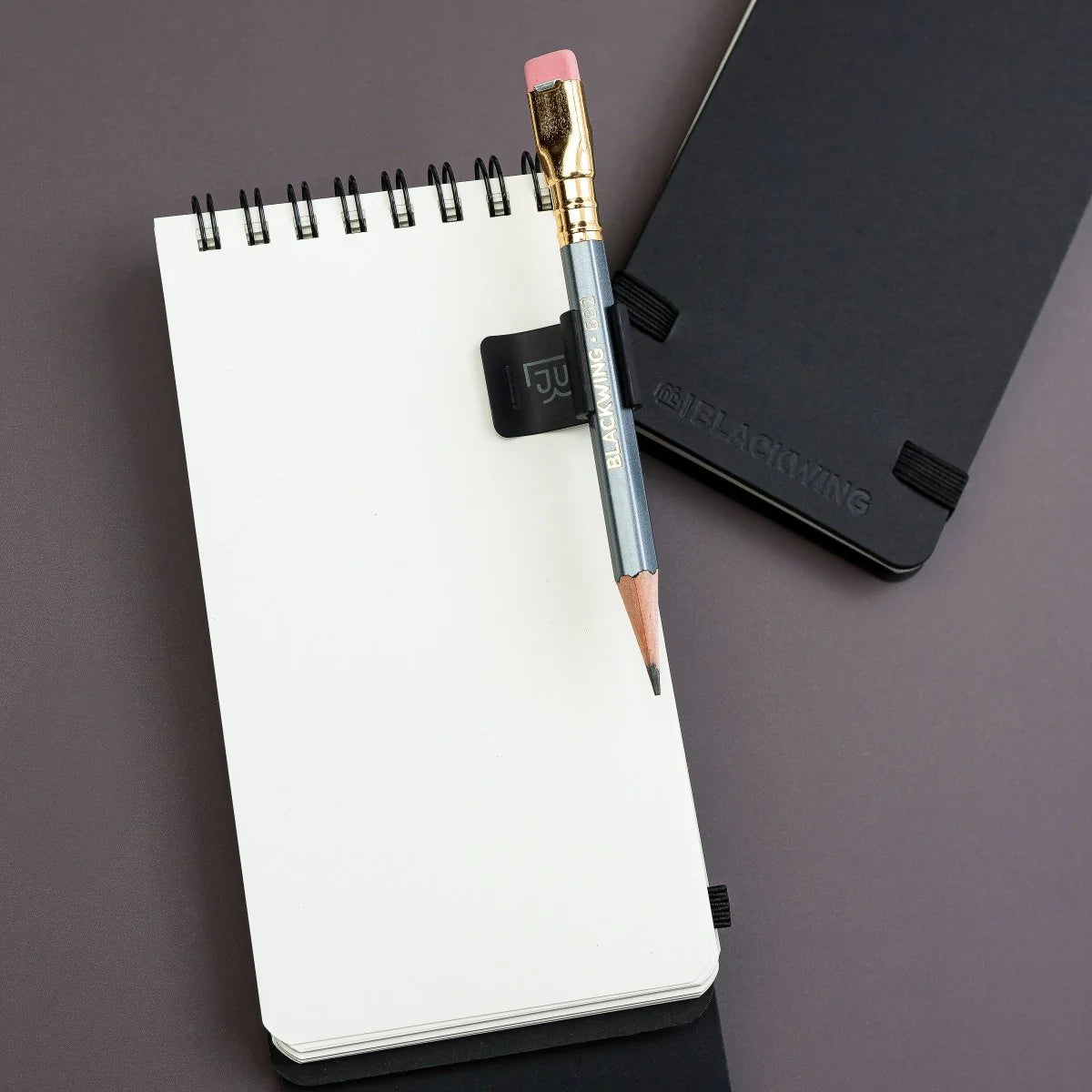 The Blackwing Notebook Clip is designed to keep your pencil and paper together. The clip is made of aluminum and finished with our signature matte black texture. Once you attach the reusable clip to your notebook, your pencil snaps into a durable thermoplastic polymer that won't scuff the lacquer. Perfect for everyday carry—clip it onto your spiral notebook, legal pad, memo book, or reading book to keep your stationery organized.