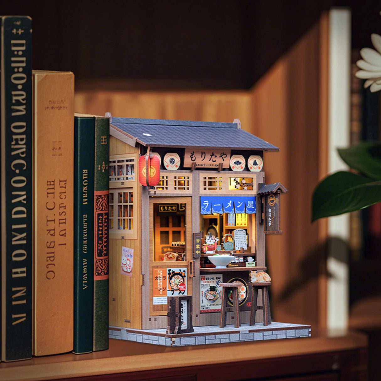From the iconic noren curtain swaying above the window to the finely detailed counter lined with ramen bowls, rice balls, and sake bottles, every element of the Ramen Sake House DIY book nook kit captures the warm, nostalgic spirit of a cozy late-night eatery tucked away in a quiet Tokyo alley. Soft LED lights bathe the scene in a gentle glow, evoking the hum of conversation, the clink of dishes, and the comforting aroma of a midnight meal.