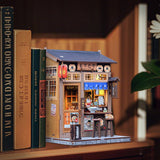 From the iconic noren curtain swaying above the window to the finely detailed counter lined with ramen bowls, rice balls, and sake bottles, every element of the Ramen Sake House DIY book nook kit captures the warm, nostalgic spirit of a cozy late-night eatery tucked away in a quiet Tokyo alley. Soft LED lights bathe the scene in a gentle glow, evoking the hum of conversation, the clink of dishes, and the comforting aroma of a midnight meal.