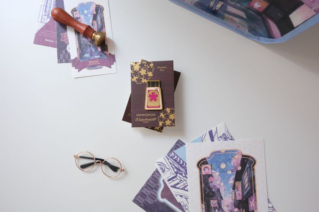The Sakura Street Ink Lapel Pin turns one of the collection's coordinating ink bottles into a collectible hard enamel design. Measuring 4.5 cm and finished with gold-plated details, the pin carries the colors and imagination of Cindy Duong's Sakura Street artwork onto a jacket, bag, or display board.