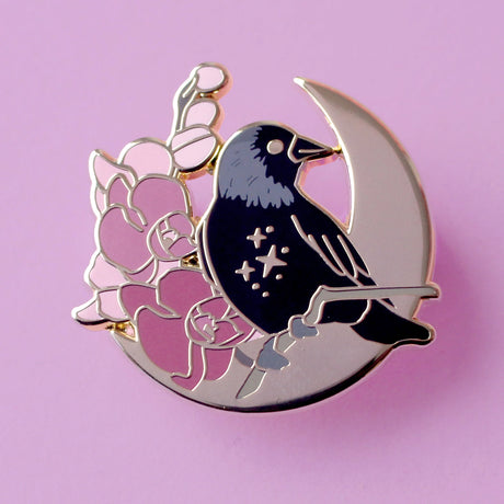 This enamel pin features a Jackdaw perching on a branch of pink orchid flowers, in front of a large gold moon. The pin is presented on a Glitter Punk backing card and is secured with 2 pin backs.

This pin is part of the Corvid Collection, an 8-part series of bird pins. 

Measurements: 35mm