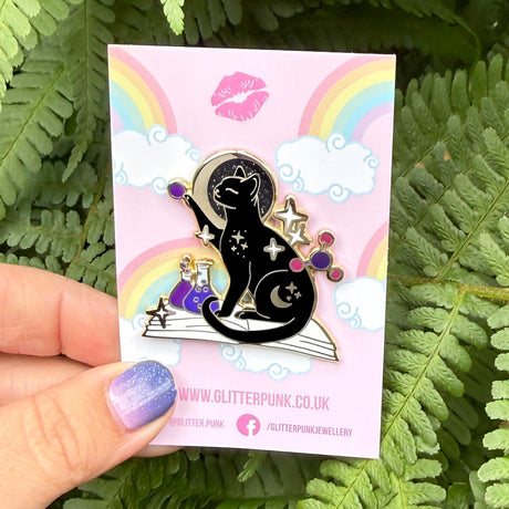 If you’re passionate about science and partial to cats, this awesome pin is perfect for you! It features a black cat sitting on a book and playing with atom parts. It also features a glittery moon and two beakers filled with purple liquid.

The pin is 42mm (1.65”) tall, and is made from gunmetal hard enamel, with glitter and UV printed details. It's secured with 2 pink clutches to keep the pin in place.