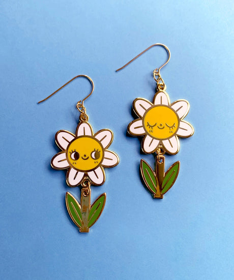 A pair of mis-matched happy daisy themed earrings (one one with happy open eyes, and one with happy closed) with separate dangling stems. Made from hard enamel with shiny gold metal finish on high quality Hypo-Allergenic, gold plated, brass based hooks.

Each earring measures approximately 5cm tall x 3cm wide. Earrings come in a Hannakin embossed gift box.