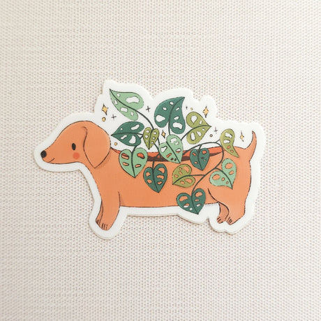 A charming dachshund planter rests among leafy greenery, a thriving cheese plant growing from its back like a small piece of playful décor. This vinyl sticker captures that cozy, plant-filled still life, adding a touch of warmth and whimsy to your everyday essentials.

Made from durable, waterproof vinyl, this sticker is designed to hold up wherever you place it—whether that’s a water bottle, laptop, or journal you reach for daily.