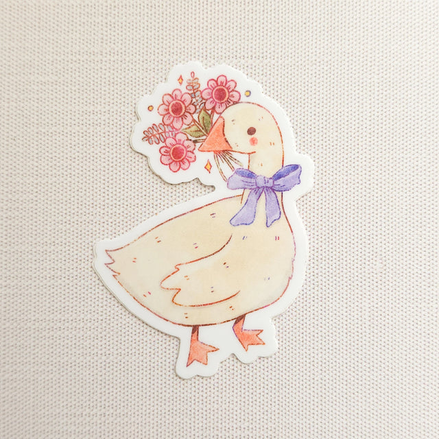 A sweet goose pauses with a bouquet of flowers held gently in its beak, a blue bow tied neatly at its neck. This vinyl sticker captures that charming, flower-filled moment, adding a warm, cottage-inspired touch to your everyday essentials.

Made from durable, waterproof vinyl, this sticker is designed to hold up wherever you place it—whether that’s a water bottle, laptop, or journal you reach for daily.