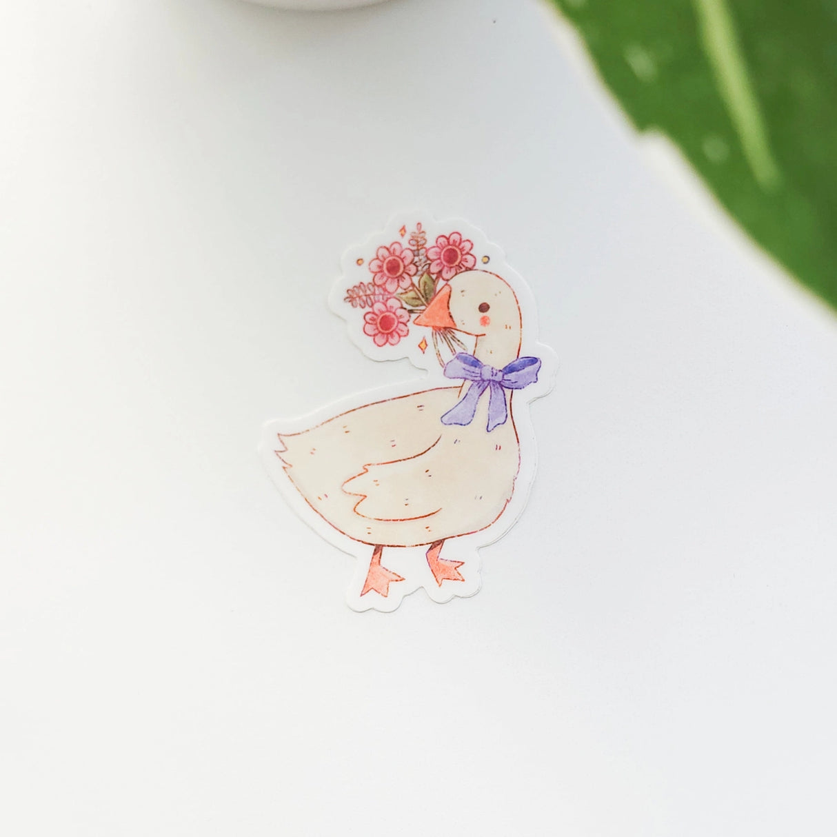 A sweet goose pauses with a bouquet of flowers held gently in its beak, a blue bow tied neatly at its neck. This vinyl sticker captures that charming, flower-filled moment, adding a warm, cottage-inspired touch to your everyday essentials.

Made from durable, waterproof vinyl, this sticker is designed to hold up wherever you place it—whether that’s a water bottle, laptop, or journal you reach for daily.