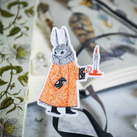 A mysterious rabbit dressed in a patterned robe carries a glowing candle through the night. This whimsical illustration blends folklore and magic, making it a perfect sticker for anyone who loves enchanting details. Printed on durable white vinyl, this sticker is waterproof, UV-proof, and made to last — ready to decorate laptops, journals, bottles, and more.
