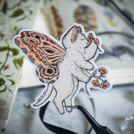 A gentle kitty with butterfly wings pauses to admire a flower, bringing a touch of magic and wonder. This enchanting design is perfect for fairy lovers, cat enthusiasts, and anyone who adores whimsical details. Printed on durable white vinyl, this sticker is waterproof, UV-proof, and long-lasting — ideal for decorating water bottles, laptops, journals, or gifts.
