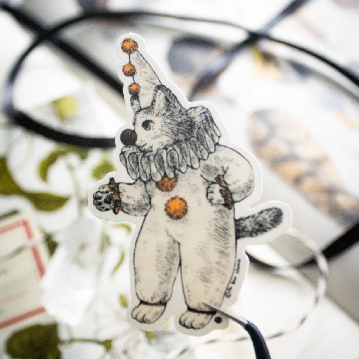 There's a kitty dressed as a clown going around looking for playful adventures. This whimsical illustration blends folklore and magic, making it a perfect sticker for anyone who loves enchanting details. Printed on durable white vinyl, this sticker is waterproof, UV-proof, and made to last — ready to decorate laptops, journals, bottles, and more.