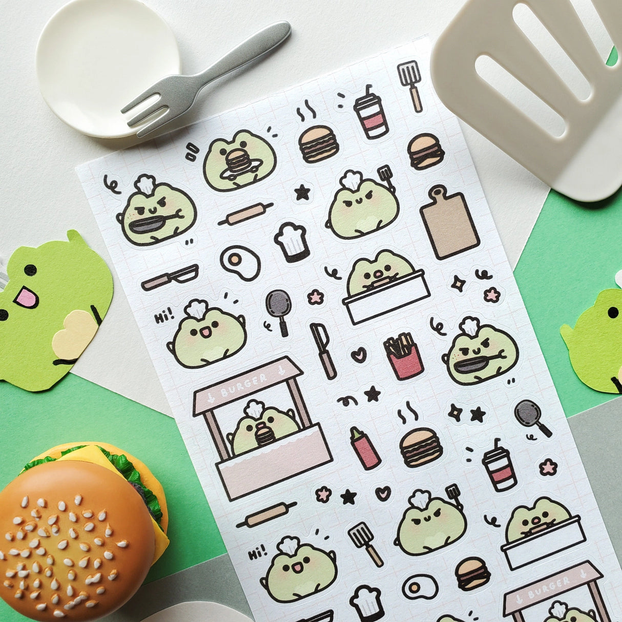 🍔🐸 Say hello to the cutest chef in town! @keziacreations’ adorable little froggy is busy in the kitchen flipping patties and serving up smiles. From sizzling burgers to crispy fries and fizzy drinks, this tiny chef is here to make your heart (and tummy) hoppy.

Art by: @keziacreations

Top Coating: None
Adhesive Layer: Paper
Backing Layer: Clear PET
6.69" x 3.54".
Weight - 0.03lbs.
1 sheet included&nbsp;