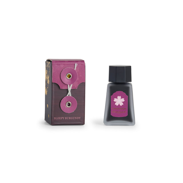 A bottle of Sleepy Burgundy ink next to its purple and brown packaging box, featuring a pink cherry blossom label, on a white background.