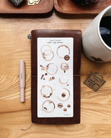 Notebook with coffee ring stickers on a wooden surface with a pen and mug.